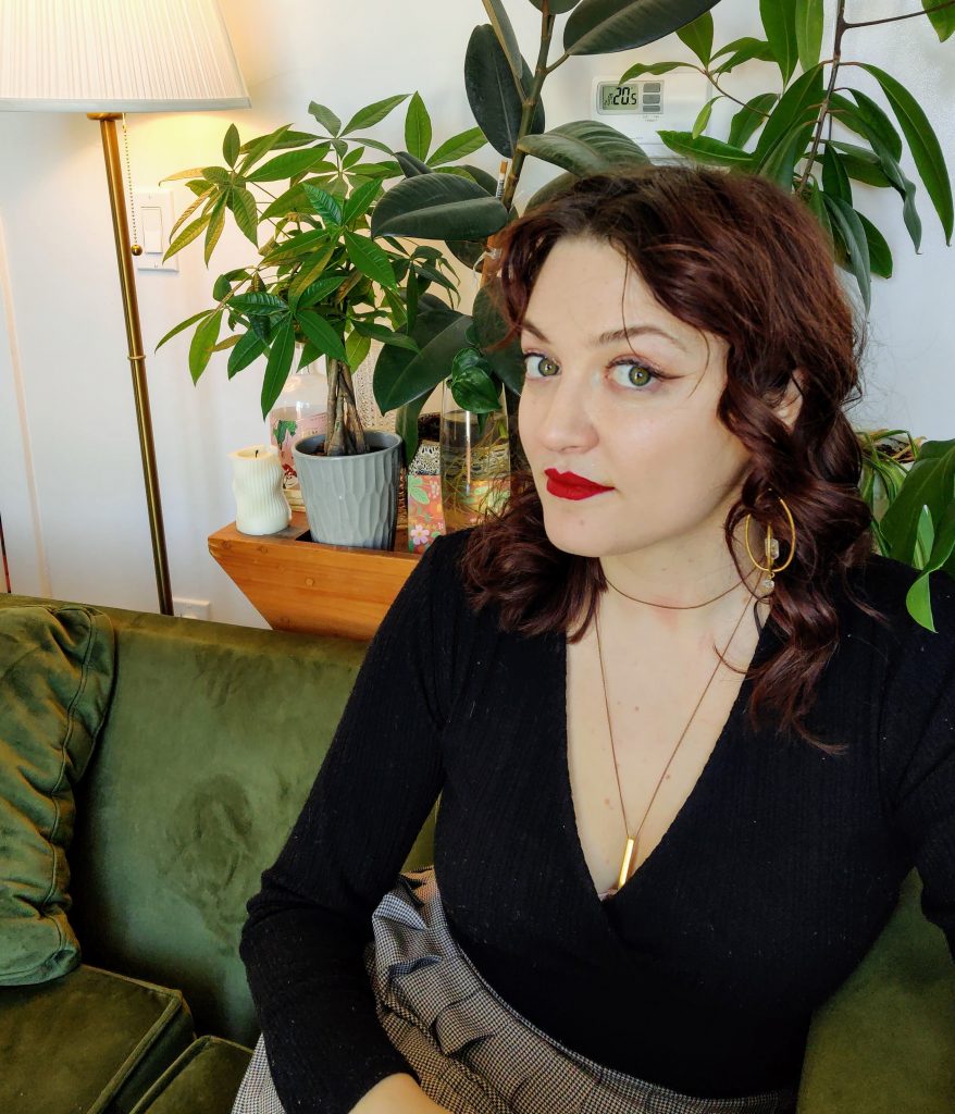 Megan looks up at the camera from her seat on a green velvet couch. She has red lipstick, curly auburn hair that goes down to her shoulders, and is wearing gold jewellery. She sits in front of numerous tropical plants. 