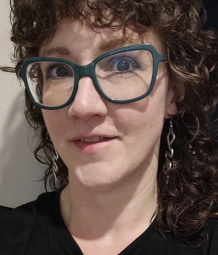 Helena smiles up at the camera. She is wearing large teal glasses and dangling silver earrings. Their brown curly hair falls to their shoulders.  