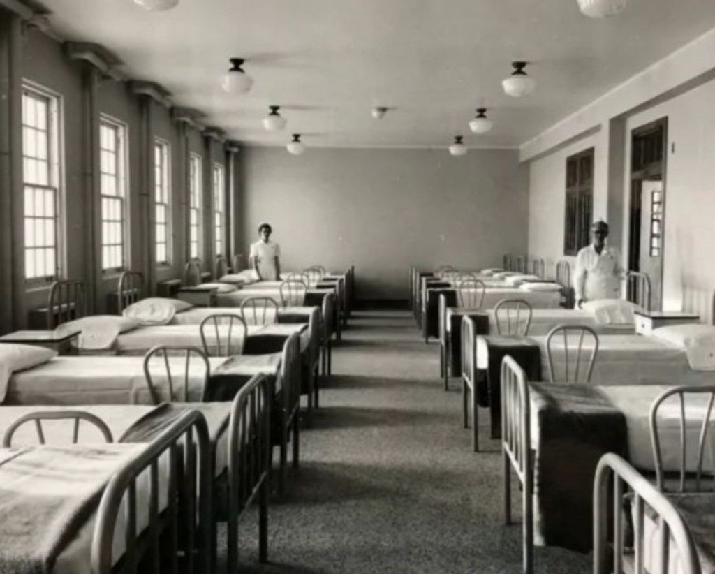 Two nurses look on in a large room full of single beds.
