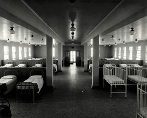 A black and white photograph of a large room with rows of single beds and cribs.